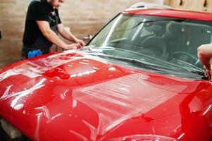 autoservicemedewerker plaatste anti-grindfilm op een rode carrosserie in de detailleringsvoertuigwerkplaats. autobescherming met speciale folies. foto