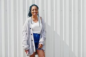 afrikaanse vrouw met dreadlocks haar, in spijkerbroek gesteld tegen witte stalen muur met mobiele telefoon in de hand. foto