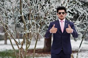 elegante Indiase macho man model op pak en roze stropdas, zonnebril geposeerd op winterdag toont duimen omhoog. foto