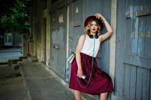 modieuze en mooie blonde model meisje in stijlvolle rood fluwelen velours rok, witte blouse en hoed, gesteld met telefoon en koptelefoon. foto