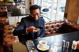 modieuze Afro-Amerikaanse man in pak en bril zit in café en snap koffie op de telefoon. foto