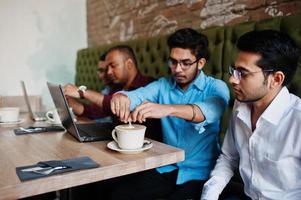 groep van vier Zuid-Aziatische mannen poseerde op zakelijke bijeenkomst in café. indianen werken samen met laptops met behulp van verschillende gadgets, praten en drinken koffie. foto