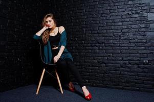 knappe brunette meisje slijtage op zwarte en groene jas in rode hoge hakken, zittend en poseren op stoel in studio tegen donkere bakstenen muur. studiomodel portret. foto