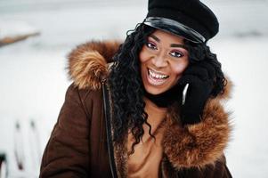 close-up gezicht van Afro-Amerikaanse vrouw in schapenvacht jas en pet gesteld op winterdag tegen besneeuwde achtergrond, met telefoon bij de hand. foto