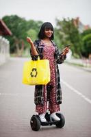 mooie afrikaanse amerikaanse vrouw die segway of hoverboard en mobiele telefoon gebruikt. zwart meisje met gele stoffen eco-tassen recycling symbool. foto