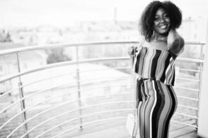 modieuze afro-amerikaanse vrouw in roze gestreepte jumpsuit, met handtas op het franse balkon. foto