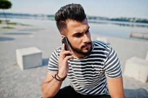 knappe lange Arabische baard man model op gestript shirt buiten geposeerd. modieuze arabische man met mobiele telefoon. foto