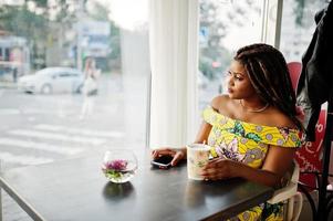 schattig klein Afrikaans Amerikaans meisje met dreadlocks, draag een gekleurde gele jurk, zit in café met een kopje koffie en kijkt naar het raam. foto