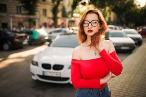 aantrekkelijke roodharige vrouw in brillen, dragen op rode blouse en jeans rok poseren op straat tegen witte sportwagen. foto