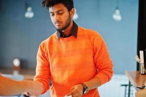 zelfverzekerde jonge indiase man in oranje trui zit aan de bar in café en berekend met de barman. foto