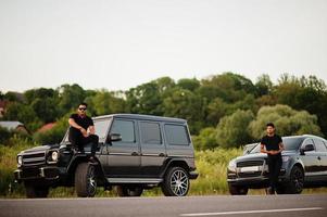 twee aziatische broers die een man dragen op alle zwarte poses in de buurt van suv-auto's. foto