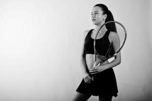 zwart-wit portret van een mooie jonge vrouw in sportkleding die een tennisracket vasthoudt terwijl hij tegen een witte achtergrond staat. foto