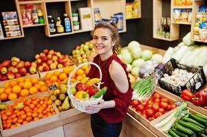 meisje in het rood met verschillende groenten en fruit in de mand in de fruitwinkel. foto