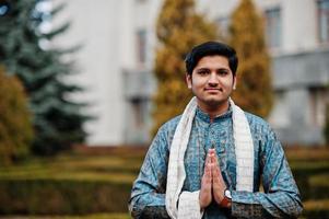 indiase man draagt traditionele kleding met witte sjaal buiten tegen groene struiken in het park, toon namaste handen teken. foto