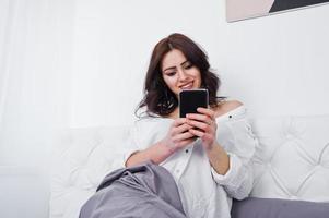 studio portret van brunette meisje in witte blouse liggend op bed en met behulp van mobiele telefoon. foto