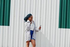 afrikaanse vrouw met dreadlocks haar, in spijkerbroek gesteld tegen witte stalen muur met mobiele telefoon in de hand. foto