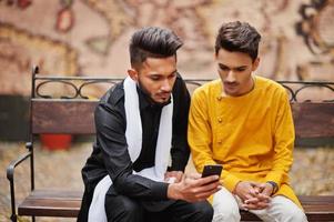 twee Indiase stijlvolle mans vrienden in traditionele kleding poseerden buiten, zittend op een bankje en kijkend naar de telefoon. foto