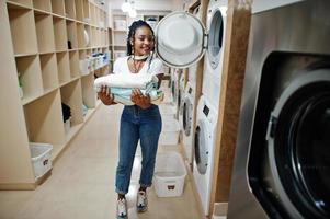 vrolijke Afro-Amerikaanse vrouw met handdoeken in handen in de buurt van wasmachine in de zelfbedieningswasserette. foto