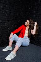 stijlvolle brunette meisje op rode jas tegen studio zwarte bakstenen muur. foto