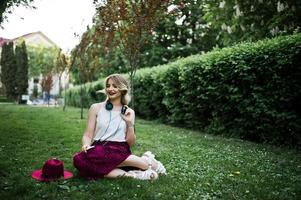 modieuze en mooie blonde model meisje in stijlvolle rood fluwelen velours rok, witte blouse en hoed, zittend op groen gras in het park met telefoon en koptelefoon. foto