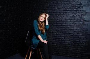 knappe brunette meisje slijtage op zwarte en groene jas in rode hoge hakken, zittend en poseren op stoel in studio tegen donkere bakstenen muur. studiomodel portret. foto