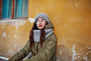 portret van brunette meisje in grijze sjaal en muts, bril bij koud weer tegen oranje muur van oud huis. foto