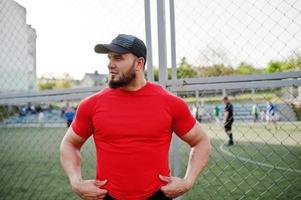 jonge brute bebaarde gespierde man draagt een rood shirt, korte broek en pet in het stadion. foto