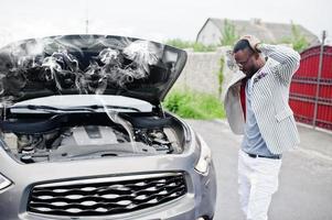 stijlvolle en rijke afro-amerikaanse man staat voor een kapotte suv-auto heeft hulp nodig bij het kijken onder geopende motorkap met rook. foto