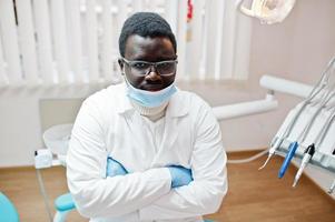 Afro-Amerikaanse mannelijke arts in masker en bril met gekruiste armen zittend op tandartsstoel in tandheelkundige kliniek.