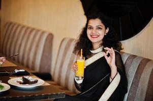 jonge indiase vrouw draagt een elegante zwarte saree zittend op restaurant met een glas sap bij de hand, ze glimlacht en toont een goed teken. foto