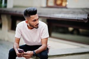 stijlvolle Indiase baard man op roze t-shirt. india model zit met mobiele telefoon bij handen buiten in de straten van de stad. foto