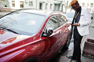 chique knappe Afro-Amerikaanse man in wit pak tegen rode luxeauto met mobiele telefoon bij de hand. foto