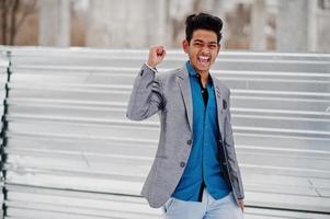 casual jonge indiase man in zilveren jasje geposeerd tegen stalen achtergrond, winnaar blije emoties tonen. foto