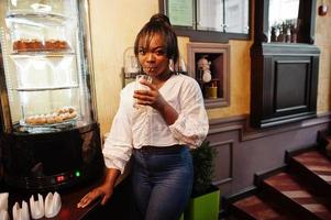 stijlvolle Afro-Amerikaanse vrouwen in witte blouse en spijkerbroek poseerden in café met karamel latte. foto