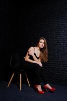 knappe brunette meisje slijtage op zwarte en rode hoge hakken, zittend en poseren op stoel in studio tegen donkere bakstenen muur. studiomodel portret. foto