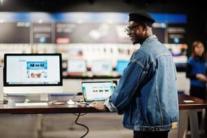 stijlvolle casual Afro-Amerikaanse man bij jeans jasje en zwarte baret met behulp van nieuwe laptop bij elektronicawinkel. foto