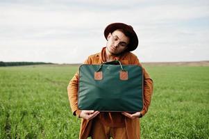 stijlvolle man met bril, bruine jas en hoed met tas op groen veld. foto