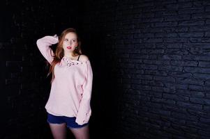 studio shot van brunette meisje op roze trui met jeans shorts tegen zwarte bakstenen muur. foto