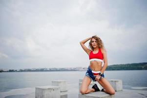 sexy krullend model meisje in rode top, jeans denim shorts en sneakers geposeerd op stenen kubussen tegen bewolkte hemel en meer. foto