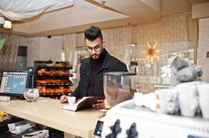 arabische man draagt een zwarte spijkerbroek en een bril in café drinkt koffie aan de bar met boek. stijlvolle en modieuze Arabische modelman. foto