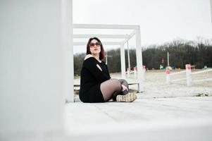 portret van brunette meisje in zwarte jurk zit op witte houten constructie. foto