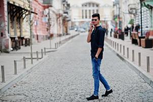 stijlvolle indiase model man in vrijetijdskleding, zwart shirt en zonnebril poseerde buiten op straat in india en sprak aan de telefoon. foto
