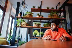 zelfverzekerde jonge indiase man in slimme vrijetijdskleding zoals een oranje trui, zittend in een café en zijn mobiele telefoon gebruikend. foto