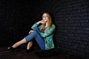 stijlvol blond meisje in jas en spijkerbroek met laptop tegen zwarte bakstenen muur in de studio. foto