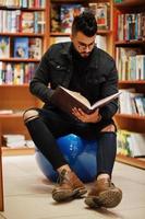 lange slimme arabische studentenman, draag een zwart spijkerjack en een bril, zit in de bibliotheek en lees een boek bij de hand. foto