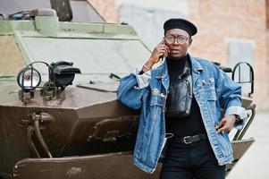 afro-amerikaanse man in spijkerjasje, baret en bril, telefonisch sprekend tegen btr militair gepantserd voertuig. foto