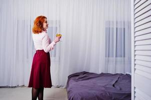 prachtig roodharig meisje in roze blouse en rode rok met glas wijn bij de hand in de buurt van bed op kamer. foto