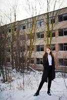 modieuze lange benen brunette model in lange zwarte mantel geposeerd buiten op winterdag tegen verlaten fabriek. foto