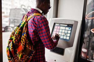 Afro-Amerikaanse man in geruit overhemd met rugzak portemonnee in de hand houden tegen ATM. zwarte man reiziger. foto