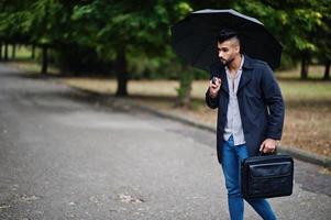 modieuze lange Arabische baard man draagt zwarte jas met paraplu en tas geval gesteld op regenweer dag. foto